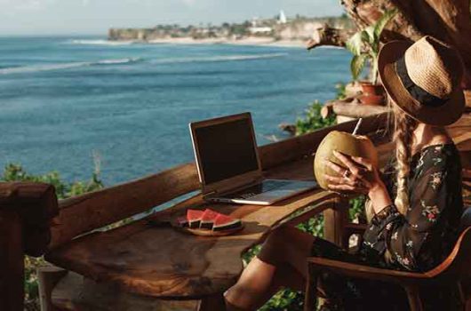 Back view young woman in straw hat sitting at table on sandy beach, enjoying fresh coconut cocktail and working on laptop